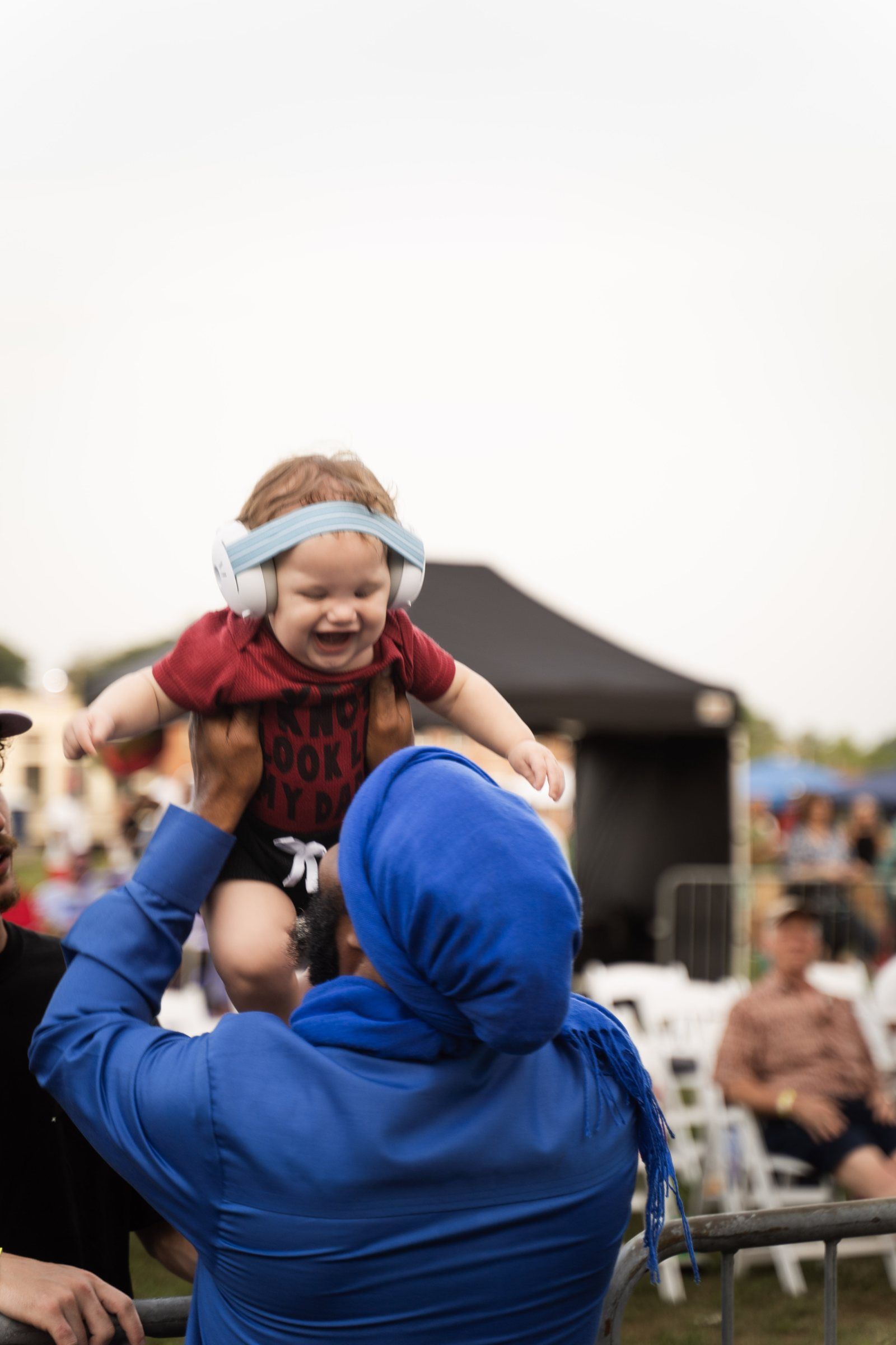 A man in a blue head wrap lifts a laughing baby wearing ear-protection earmuffs above the crowd, 2024